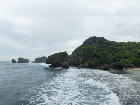 Aerial view of Siung Beach with dramatic limestone cliffs, lush greenery, and a suspension bridge connecting rock formations over crashing waves at Tepus, Special Region of Yogyakarta, Indonesia.