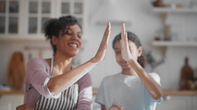 Pretty black woman teaching her daughter to cook at home, hitting palms, smiling . Family time in weekend, give five for success, charming teen girl and her mother cooking together from family diner