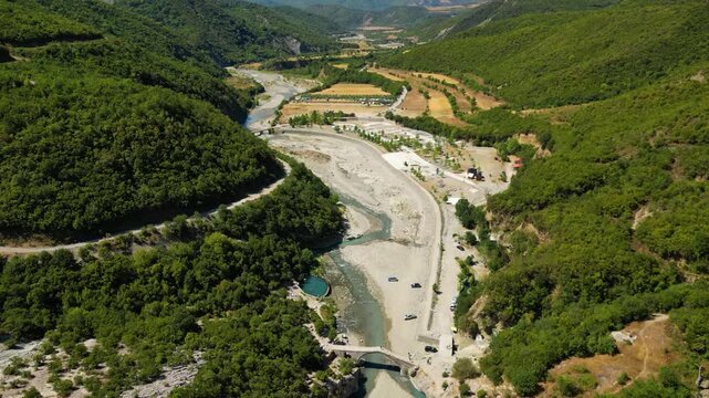 Aerial view of the historic Ottoman Katiu Bridge and circular turquoise thermal baths at the entrance of Langarica Canyon near Permet, Albania, set against the backdrop of the rugged Nemercka
