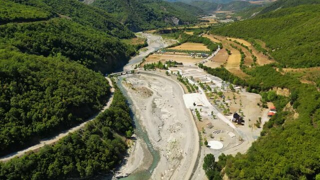 Aerial view of the historic Ottoman Katiu Bridge and circular turquoise thermal baths at the entrance of Langarica Canyon near Permet, Albania, set against the backdrop of the rugged Nemercka