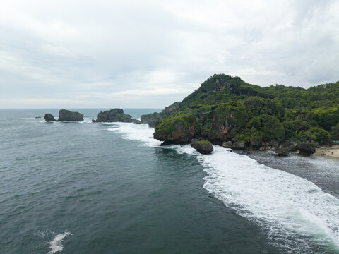Aerial view of Siung Beach featuring dramatic limestone cliffs, lush green hills, and white waves crashing against the rocky coastline in Tepus, Special Region of Yogyakarta, Indonesia.