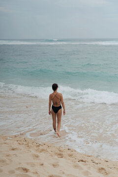 Woman in strappy bikini walking toward ocean waves, Uluwatu, Bali