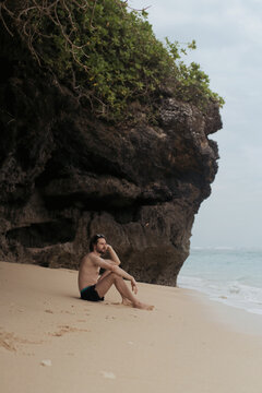 Pensive young man sitting on beach beside limestone cliff, Bali
