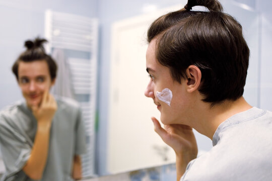 Young person applying facial cream while looking in bathroom mirror