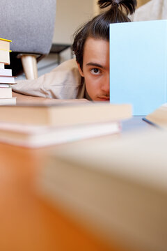 Curious adolescent peeking over book cover while surrounded by books