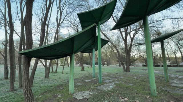 Large green metal structures rise above a mosaic stone path on a chilly morning. The frost-covered grass and bare trees create a quiet, mysterious atmosphere in this peaceful park landscape.
