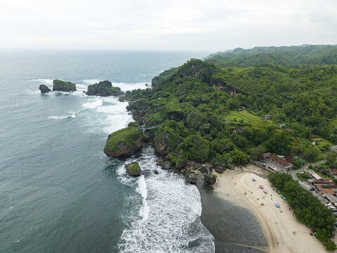 Aerial view of Siung Beach featuring dramatic rocky cliffs, lush green hills, and white sand meeting the ocean waves in Tepus, Special Region of Yogyakarta, Indonesia.