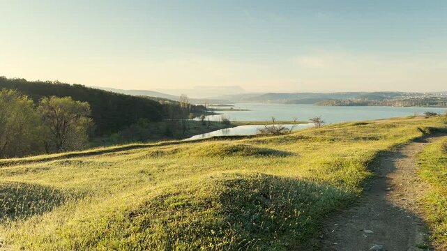 Grassy hills roll gently toward a wide blue lake during a quiet morning. This vast landscape feels peaceful and free. The soft sunlight on the field creates a sense of pure relaxation.