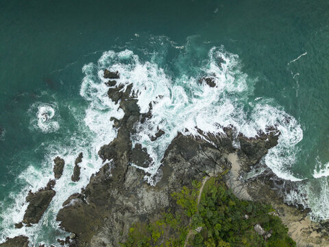 Aerial view of Siung Beach with turquoise ocean waves crashing against dark volcanic rocks and lush green trees in Tepus, Special Region of Yogyakarta, Indonesia.