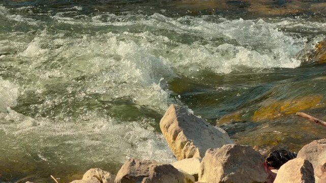 Cold mountain water crashes and foams against jagged shore rocks. The powerful, energetic flow creates a sense of wild freedom and natural strength. Sunlight reflects off the churning spray.
