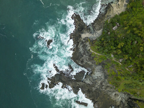 Aerial view of Siung Beach with turquoise waves crashing against dark jagged rocks and lush green vegetation in Tepus, Special Region of Yogyakarta, Indonesia.
