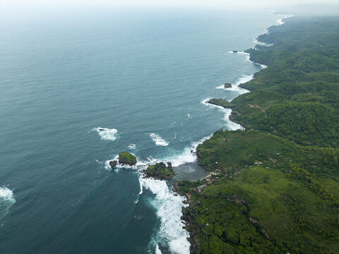 Aerial view of Siung Beach featuring rugged limestone cliffs, turquoise ocean waves crashing against rock formations, and lush green hills in Tepus, Special Region of Yogyakarta, Indonesia.