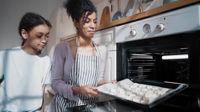 Bake tasty homemade cookies, black woman and her daughter using oven for baking. African american family, mother and her teen child admiring cookies on baking sheet, cooking delicious sweets at home