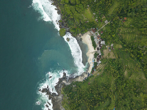 Aerial view of Siung Beach with white sand, turquoise waves crashing against rocky cliffs, and lush green tropical vegetation in Tepus, Special Region of Yogyakarta, Indonesia.