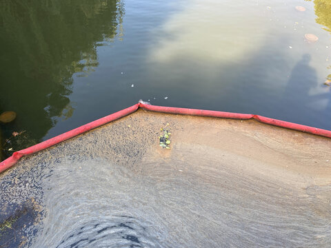 Red floating barrier blocking industrial foam and debris from spreading in waterway. Environmental protection equipment installed near concrete pipe outlet to prevent water contamination.
