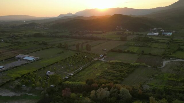 Aerial panorama of a small, green eco-campsite nestled along the banks of the Buna River in Northern Albania, featuring lush agricultural fields and mountain views under a soft sunset sky.