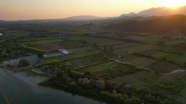 Aerial panorama of a small, green eco-campsite nestled along the banks of the Buna River in Northern Albania, featuring lush agricultural fields and mountain views under a soft sunset sky.