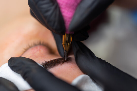 Close up of permanent eyebrow makeup procedure, beautician performing microblading with precision tool in beauty salon