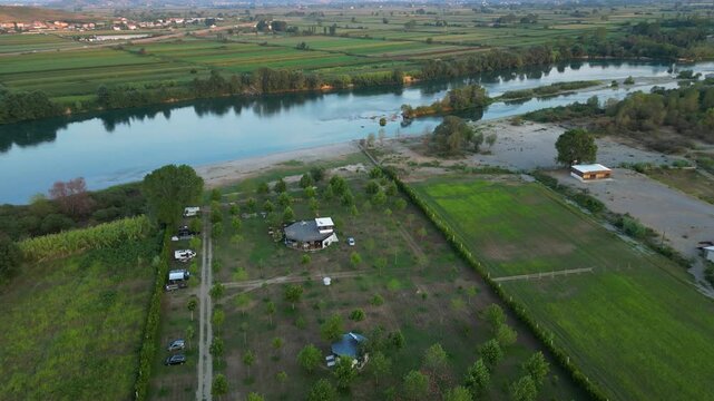 Aerial panorama of a small, green eco-campsite nestled along the banks of the Buna River in Northern Albania, featuring lush agricultural fields and mountain views under a soft sunset sky.
