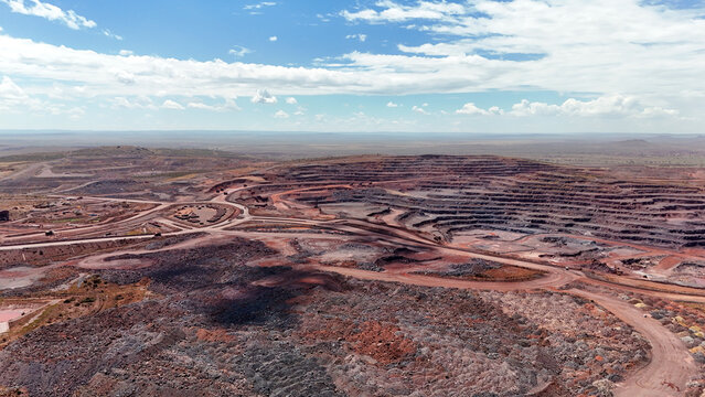 Aerial view of the vast, arid landscape scarred by the geometric patterns of a mining operation, its layers of rust-red earth contrasting with the pale sky, North West, South Africa.