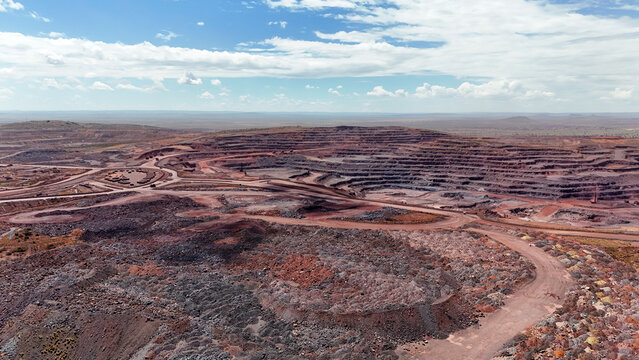 Aerial view of a vast, scarred open-pit mine with deep red earth and dark terraced pits contrasting with the bright sky, Rustenburg, North West, South Africa.