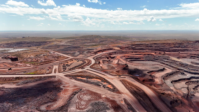 Aerial view of the sprawling, scarred landscape of an open-pit mine, where deep red earth contrasts sharply with the pale blue sky and distant horizons, Rustenburg, North West, South Africa.