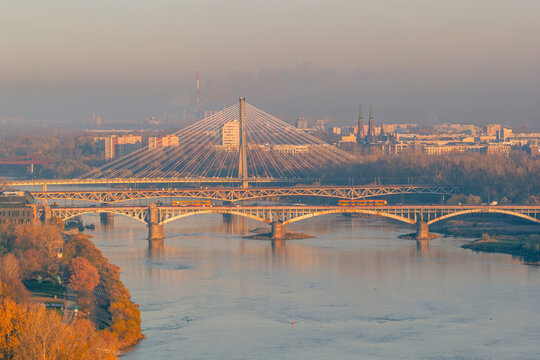 Aerial view of Swietokrzyski Bridge and Poniatowski Bridge spanning the Vistula River with autumn trees and city skyline in golden hour light Warsaw, Masovian Voivodeship, Poland.