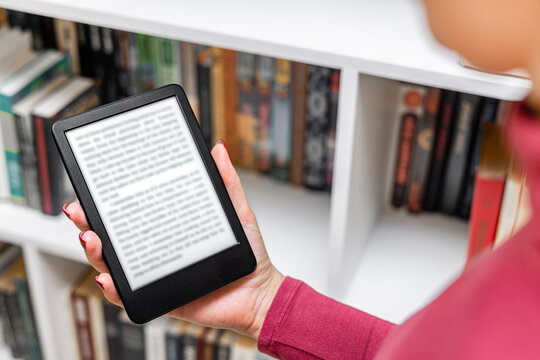 A woman reads an e-reader near a bookcase. an e-book against the background of paper books. ease of use of an e-book. a modern way to read books. 