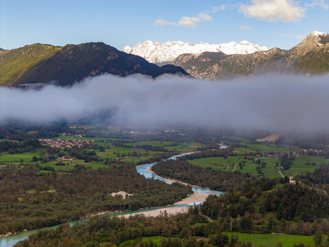 Aerial view of the Soca River winding through a lush green valley with low-lying clouds and snow-capped mountain peaks in Srpenica, Tolmin, Slovenia.