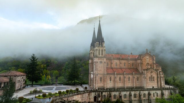 Aerial view of the Basilica's spires pierce the mist-laden mountains, a symphony of stone and nature's breath, Basilica de Santa Maria la Real de Covadonga, Cangas de Onis, Spain.
