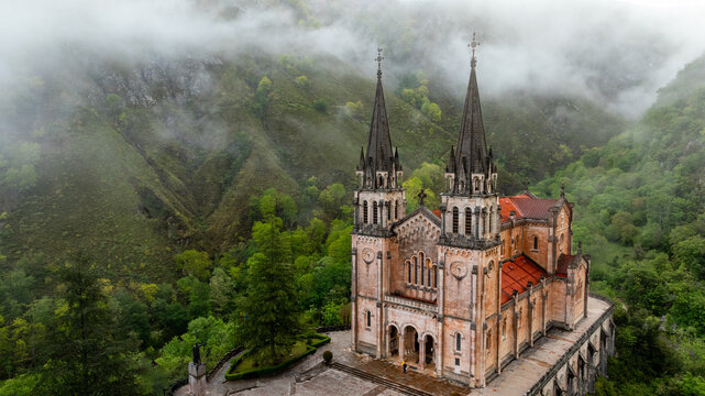 Aerial view of the grand basilica stands majestically against a backdrop of mist-shrouded mountains and verdant forests, its spires reaching towards the heavens, Cangas de Onis, Spain.