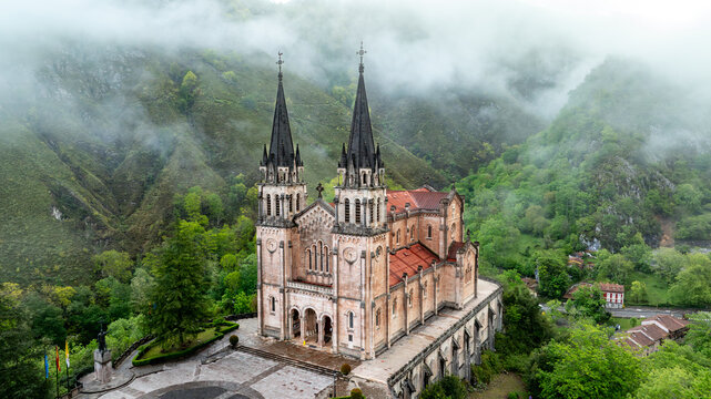 Aerial view of Basilica de Santa Maria la Real de Covadonga, a symphony of stone amidst verdant hills veiled in ethereal mist, Cangas de Onis, Spain.