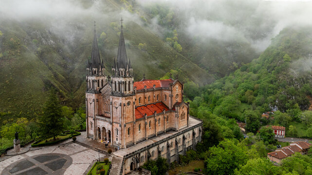 Aerial view of the Basilica de Santa Maria la Real de Covadonga rises majestically amidst verdant hills shrouded in ethereal mist, its spires piercing the heavens, Cangas de Onis, Spain.