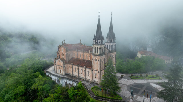 Aerial view of the Basilica's spires pierce through the swirling mist, contrasting against the verdant landscape, Basilica de Santa Maria la Real de Covadonga, Cangas de Onis, Spain.
