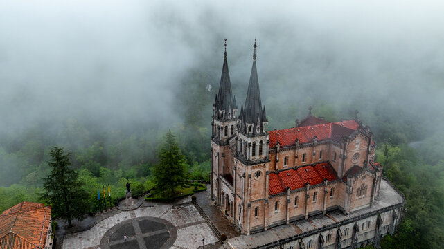 Aerial view of the Basilica of Santa Maria la Real de Covadonga, its spires piercing through the mist-laden peaks, a striking contrast of stone and verdant forest, Cangas de Onis, Spain.