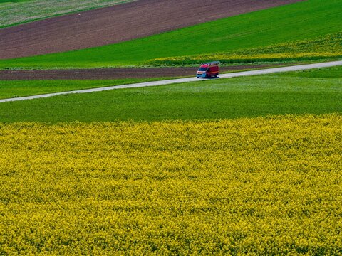 Aerial view of a red truck on road, amidst bright yellow, green, and brown fields, Wintzenheim-Kochersberg, Grand Est, France.