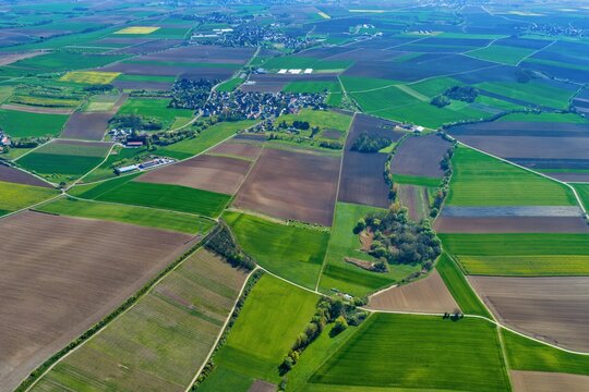 Aerial view of a patchwork of vibrant green and rich brown agricultural fields encircling a tranquil village under a clear sky, Wintzenheim-Kochersberg, Grand Est, France.