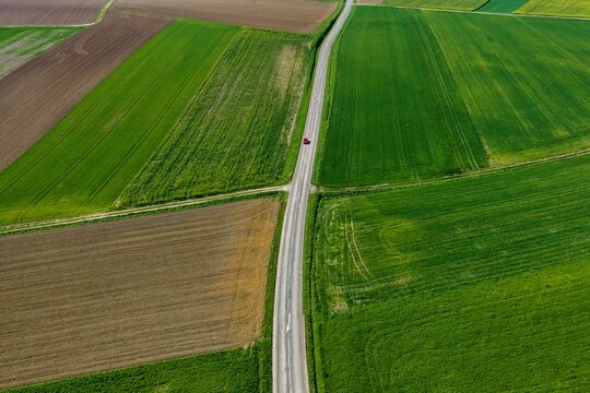 Aerial view of vibrant green and brown agricultural fields, bisected by a long road and a red car, Wintzenheim-Kochersberg, Grand Est, France.