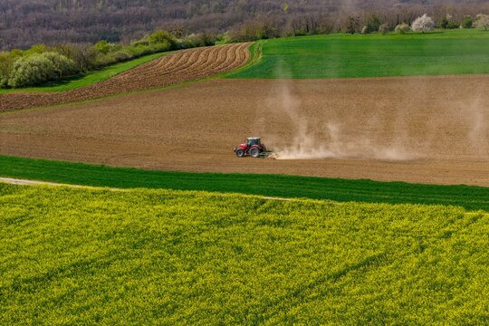 Aerial view of a red tractor tilling brown earth, bordered by vibrant yellow fields and distant green hills, Wintzenheim-Kochersberg, Grand Est, France.