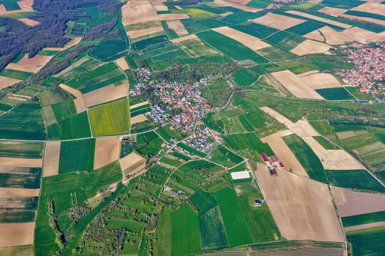 Aerial view of a vibrant patchwork of green and brown agricultural fields encircling a charming village with reddish-orange rooftops, Wintzenheim-Kochersberg, Grand Est, France.