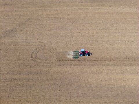 Aerial view of a vibrant red and blue tractor tilling a vast, muted brown field, leaving a dusty trail, Wintzenheim-Kochersberg, Grand Est, France.