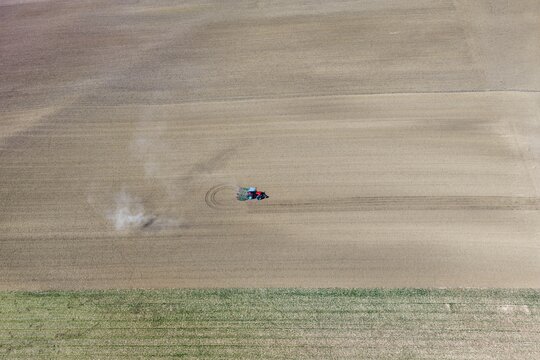 Aerial view of a tractor tilling dry, pale earth, creating dust plumes, with a sharp contrast to a green field below, Wintzenheim-Kochersberg, Grand Est, France.
