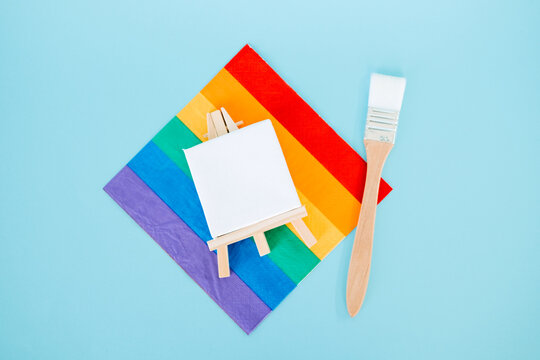 Small blank canvas on easel rests on rainbow pride flag napkin next to paint brush. LGBTQ mockup.Artistic social movement, diversity in arts, creative expression freedom