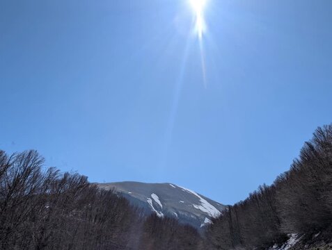   
Paesaggio montano con pendii boscosi in ombra e una vetta pi&ugrave; alta illuminata dal sole, con tracce di neve e cielo limpido. Un&rsquo;atmosfera luminosa e naturale