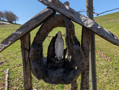   
Piccolo santuario montan con statua votiva protetta da una struttura in legno, immerso in un paesaggio naturale con prato e cielo limpido. Una scena che richiama spiritualit&agrave;, tradizione e quiete