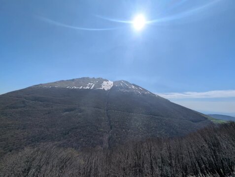   
Paesaggio montano con pendii boscosi in ombra e una vetta pi&ugrave; alta illuminata dal sole, con tracce di neve e cielo limpido. Un&rsquo;atmosfera luminosa e naturale
