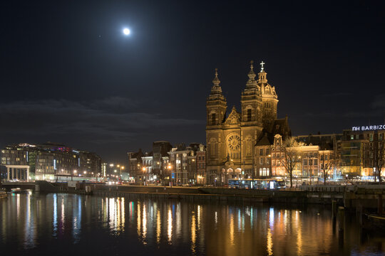 View of the illuminated facade of the Basilica of Saint Nicholas mirrored in the still waters under a bright moon, Amsterdam, North Holland, Netherlands.