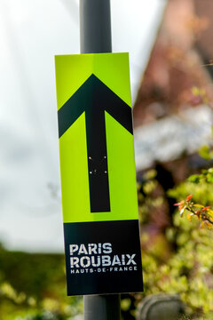 Green directional sign with black arrow for the Paris Roubaix cycling race, mounted on a pole, guiding route in an outdoor setting.