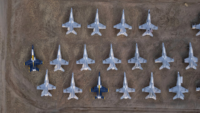 Tucson, United States - 30 January 2025: Aerial view of rows of grey fighter jets on dry desert ground, two strikingly colored planes breaking the pattern.