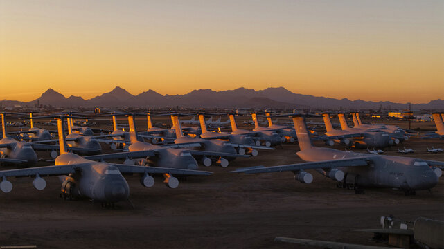 Tucson, United States - 06 February 2025: Aerial view of hulking grey aircraft on a dusty field under a fiery sunset, framed by dark mountains.